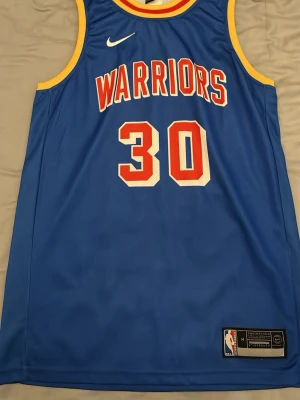 Blå Warriors basketlinne Curry #30 - Snyggt blått Golden State Warriors basketlinne från Nike. tröjan är lite retro och väldigt skön att ha på sommaren när det är varmt och skön att träna i. Storleken är M