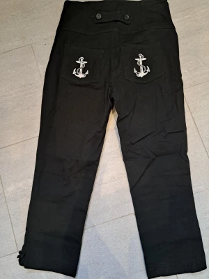 Svarta sailorbyxor med ankardetaljer - Unika svarta sailorbyxor med ankare broderade på bakfickorna. Byxorna har hög midja, raka ben och dekorativa knappar både fram och bak. Tillverkade i ett kraftigare bomullstyg som ger en klassisk marin känsla. Slutar på vaden. Använda en gång. Annars helt nya och inget att anmärka på 