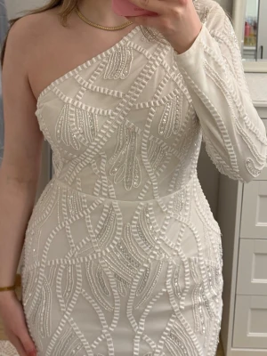 Vit one shoulder klänning med pärlor - Snygg vit klänning med one shoulder-design och glittriga pärldetaljer. Klänningen har figurnära passform och ett unikt mönster av pärlor och paljetter över hela tyget. Perfekt för dig som vill sticka ut med en elegant look. Använd en ggn!!