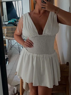 Vit kort klänning med smock - Supersöt vit kort klänning med smockad midja och djup v-ringning. Klänningen har knytband på axlarna och ett flowy, volangigt kjolparti. Perfekt för varma dagar och ger en fräsch och trendig look.