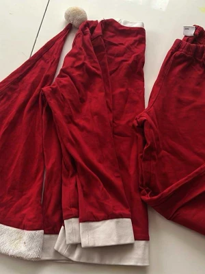 Rött tomte-set med långärmad tröja - Säljer ett rött tomte-set med långärmad tröja, byxor och matchande tomteluva. Setet har vita detaljer vid ärmslut, benslut och halsringning samt en vit fluffig kant på luvan. Perfekt för julens mysiga stunder. Materialet är mjukt bomullstyg.