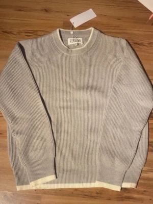 Maison Margiela - Säljer en stilren grå ribbstickad tröja från Maison Margiela. Tröjan har rund halsringning och kontrasterande vita detaljer vid hals, ärmslut och nederkant. Perfekt för dig som gillar minimalistisk design och exklusiva material.