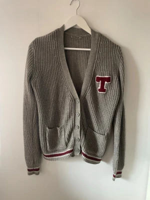 Grå kofta från Terranova med T-detalj - Säljer en grå ribbstickad kofta från Terranova med knappar framtill, två fickor och en rödvit T-applikation på bröstet. Koftan har långa ärmar och rödvita ränder vid ärmslut och nederkant. Står att det är storlek L men skulle säga att det är mer en M.