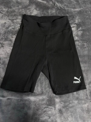 Svarta ribbade shorts från Puma - Säljer ett par svarta ribbade shorts från Puma med hög midja och logga på benet. De är stretchiga och perfekta för träning eller chill. Klassisk biker-modell med bekväm passform.