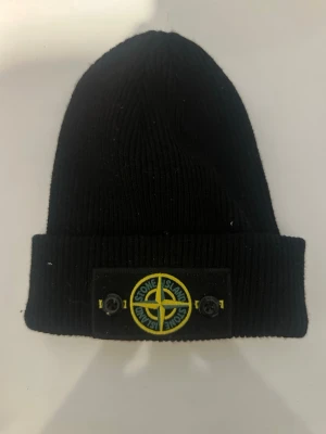 Svart mössa från Stone Island - Säljer en svart ribbstickad mössa från Stone Island med uppvikt kant. Mössan har den klassiska Stone Island-loggan framtill i gult och grönt samt en vit etikett med märkesnamnet på baksidan. Perfekt accessoar för dig som gillar streetwear och vill ha en stilren look. Pris går ner vid snabb affär 