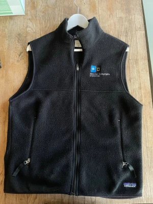 Svart fleeceväst Patagonia x NatGeo - Svart fleeceväst från Patagonia med broderad National Geographic Endeavour-logga på bröstet. Västen har hel dragkedja framtill, två fickor med dragkedja och hög krage. Perfekt för lager-på-lager och outdoor-vibe.