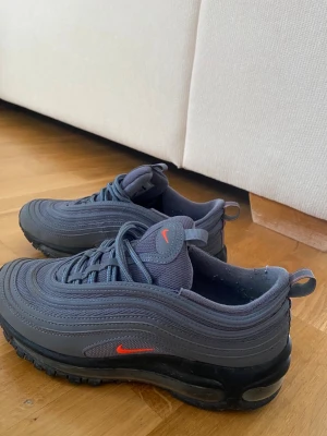Nike Air Max 97 mörkgrå sneakers - Nike Air Max 97 sneakers i mörkgrått med vågformade detaljer och en röd Swoosh-logga på sidan och plösen. Skorna har en synlig Air-sula och är tillverkade i syntetmaterial och mesh för en sportig look.