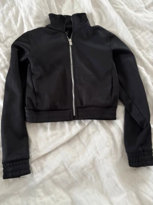 Svart croppad träningsjacka - Snygg svart croppad träningsjacka med dragkedja framtill och ribbade muddar vid ärmslut och midja. Jackan har en stilren design och passar perfekt till streetwear eller en chill vardagslook.🌸 Köpt för 299💓