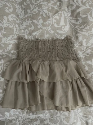 Beige volangkjol från Neo Noir - Supersöt beige kjol från Neo Noir med bred smock i midjan och dubbla volanglager. Kjolen har en luftig och flowy känsla, perfekt för dig som gillar en feminin och trendig stil. Materialet känns lätt och bekvämt mot huden.