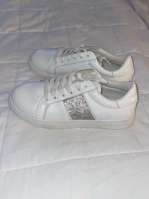 Vita sneakers med glitterdetalj - Snygga vita sneakers med silvrig glitterdetalj på sidan. Skorna har klassisk låg profil, vita snören och är tillverkade i ett slätt material som ser ut som konstläder. Perfekta för dig som vill ha en clean look med lite extra bling.
