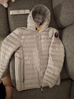 Ljusgrå dunväst från Parajumpers - Snygg ljusgrå dunväst från Parajumpers med huva och gul detalj vid halsen. Västen har quiltad design, dragkedja framtill och flera fickor. Märkespatch på axeln och mörk insida. Perfekt för dig som vill ha både stil och funktion. Äkta wallah