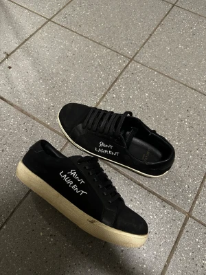 Svarta sneakers från Saint Laurent - Svarta sneakers från Saint Laurent med vit logotyptext på sidan. Skorna har svarta skosnören, vit sula och är tillverkade i en kombination av läder och textil. Klassisk låg modell med stilren design och diskreta detaljer. Priset är ej hugget i sten så varm inte rädda att ställa frågor 😁