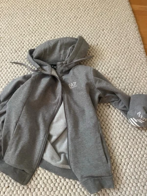 Grå hoodie från EA7 Emporio Armani - Snygg grå hoodie från EA7 Emporio Armani med dragkedja framtill och huva med dragsko. Tillverkad i mjukt material med borstad insida för extra komfort. EA7-logga på bröstet och ärmen ger en sportig vibe. Perfekt till chill eller streetwear.