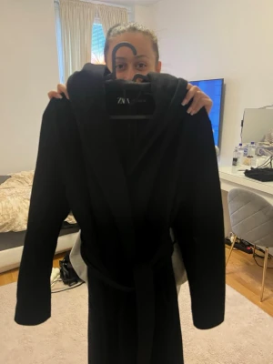 Svart morgonrock med huva från ZARA - Kappa i svart färg från ZARA med huva och knytskärp i midjan. Kappan har långa ärmar och är gjord i ett mjukt material som passar perfekt för utgång. Enkel och stilren design som känns modern.