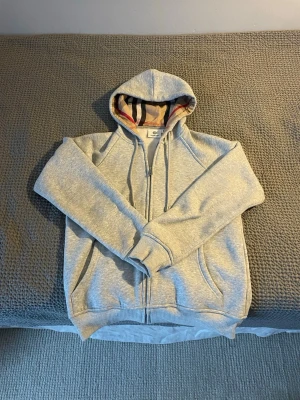 Grå zip hoodie från Burberry - Snygg grå zip hoodie från Burberry med klassiskt rutigt foder i huvan. Hoodien har dragkedja framtill, två fickor och ribbade muddar vid ärmar och nederkant. Perfekt för dig som gillar stilrena plagg med exklusiv känsla.