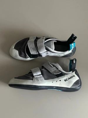 Scarpa klätterskor med kardborre - Säljer ett par svarta och gråa klätterskor från Scarpa med vita och svarta detaljer.🧗🧗‍♀️ Skorna har två breda kardborreband och slitstark Vibram-sula. Perfekta för dig som vill ha bra grepp och snygg design på klätterväggen.                               ✅Pris kan diskuteras