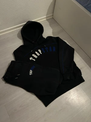 Svarta TrapStar tracksuit - Säljer ett par svarta tracksuit från Trapstar med blå och vita detaljer samt broderad logga på benet. Byxorna har resår i midjan och benslut samt dragskor för bästa passform. Perfekta för chill dagar och streetwear-stil. Finns fler bilder ;) (Storlek L)