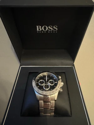 Hugo Boss silverfärgad klocka - Stilren klocka från Hugo Boss med svart urtavla och tre mindre subdials. Klockan har en robust länk i silverfärgat metall och tydliga markeringar runt boetten. Perfekt accessoar för dig som gillar klassisk och modern stil. Länkar tillkommer för att justera strl och orginal lådan. Nypris 1999kr pris går att diskutera vi snabb affär.