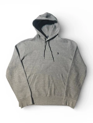 Grå hoodie från Polo Ralph Lauren | M - Klassisk grå hoodie från Polo Ralph Lauren med svart broderad logga på bröstet. Hoodien har känguruficka, svarta dragsnören och ribbade muddar. Tillverkad i mjukt bomullsmaterial för en skön och avslappnad stil.