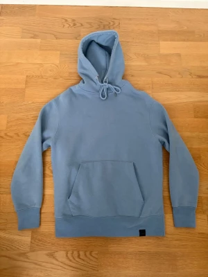 Ljusblå hoodie med magficka  - Säljer en stilren ljusblå hoodie med huva och dragsko från PULL & BEAR. Hoodien har en stor magficka framtill och ribbade muddar vid ärmslut och nederkant. Perfekt för dig som gillar en enkel och clean look. Materialet känns mjukt och skönt mot huden. Storlek XS men sitter som en S. Pris kan alltid diskuteras✅.