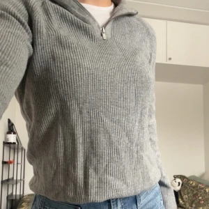 Grå ribbstickad quarter zip - Säljer en stilren grå ribbstickad tröja med halv dragkedja i halsen/quarter zip-stil. Tröjan har lång ärm och en avslappnad passform. I botten har det på två ställen gått upp lite (se bild), men har använt tröjan flera gånger utan att det går upp mer. Döljs enkelt genom att vika upp tröjan lite