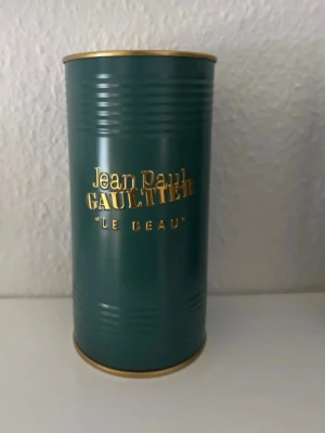 Jean Paul Gaultier Le Beau parfym - Snygg parfym från Jean Paul Gaultier, Le Beau, i en mörkgrön cylinderformad metallförpackning med guldfärgade detaljer och text. Förpackningen har en modern och lyxig känsla som sticker ut på hyllan.