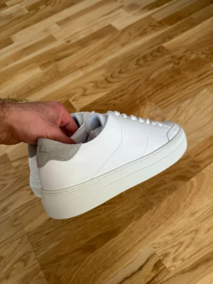 Vita sneakers från J.Lindeberg - J.Lindeberg vita sneakers – helt nya, aldrig använda. Säljs utan kartong. Stilren design med grå mockadetalj baktill. Bekväma och lättmatchade till både vardag och uppklätt.  Nypris 2000