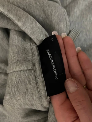 Grå hoodie Peak Performance XS - Säljer en ljusgrå hoodie från Peak Performance i storlek XS. Hoodien har stor huva med dragsko, magficka och svart tryck med logga på bröstet. Tillverkad i mjukt bomullsmaterial och har ribbade muddar vid ärmar och nederkant. 