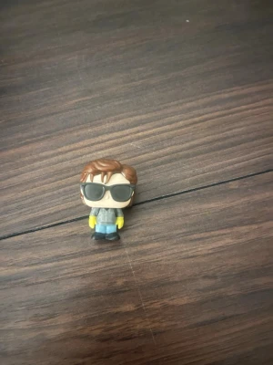 Steve stranger things kinder joy  - Säljer en liten vinylfigur med brunt hår, stora svarta solglasögon, grå jacka, blå byxor och gula handskar. Figuren har en stilren och lekfull design och passar perfekt som dekoration eller samlarobjekt.