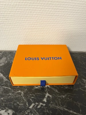 Svart plånbok  - Snygg svart plånbok med klassiskt monogrammönster i grått. Tillverkad i läder och levereras i original orange ask. Perfekt för dig som vill ha en stilren och lyxig accessoar.