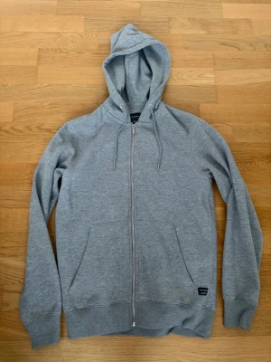 Grå zip-hoodie med fickor - En stilren grå hoodie med dragkedja framtill, huva med dragsko och två stora fickor från Jack & Jones. Perfekt för lager-på-lager och chill dagar. Mjuk bomullsblandning och ribbade muddar vid ärmslut och nederkant. Storlek S. Pris kan alltid diskuteras✅. 