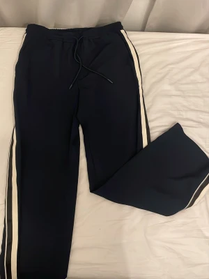 Svarta mjukisbyxor från ZARA - Säljer ett par svarta mjukisbyxor (ZARA) med vita sidoremsor och dragsko i midjan. Byxorna har en avslappnad passform och är perfekta för chill dagar. Materialet känns mjukt och bekvämt, och designen är enkel men ändå snygg.