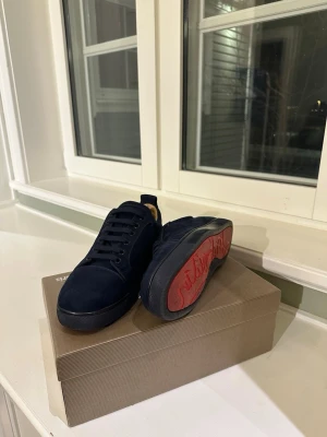 Mörkblå sneakers från Christian Louboutin - Snygga mörkblå sneakers i mocka från Christian Louboutin med klassisk röd sula och diskret logga på hälen. Skorna har mörka snören och en låg profil som ger en clean och stilren look. Perfekta för dig som gillar exklusiva detaljer och streetstyle.