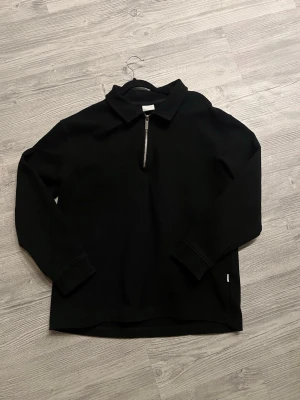 Svart långärmad tröja med dragkedja - En stilren svart långärmad tröja med krage och halv dragkedja framtill. Tröjan har en avslappnad passform och är tillverkad i ett mjukt material som passar perfekt till många olika outfits. Enkel och clean design utan synliga loggor.