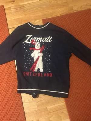 Mörkblå stickad tröja Zermatt Hollister - Mörkblå stickad tröja från Hollister med avslappnad passform. Stor vit isbjörn med röd halsduk och skidor samt texten 'Zermatt Switzerland' på framsidan. Vita och röda detaljer, perfekt för vintriga vibes.