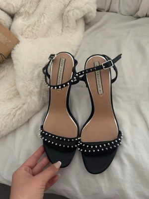 Svarta sandaler med nitar från Zara - Snygga svarta sandaler från Zara Trafaluc med tunna remmar och silverfärgade nitar längs framremmen. Öppen tå och justerbart spänne vid ankeln. Klack i låg modell och stilren design som passar till många outfits. Dom passa de som bär 39,5 och 40 för den är ganska små