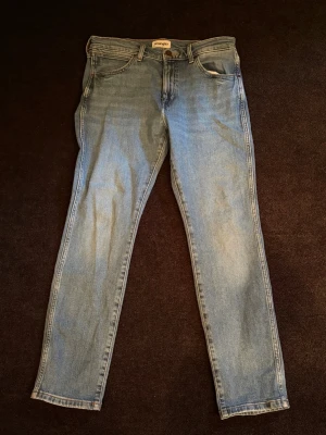 Ljusblå jeans från Wrangler - Säljer ett par klassiska ljusblå jeans från Wrangler med slim fit och fem fickor. Jeansen har en snygg tvätt och detaljer som Wrangler-lappen bak i midjan. Använd endast ett fåtal gånger. Nypris ca 900, storlek 30/30