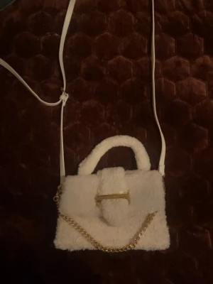 Vit fluffig handväska med guldkedja från Aldo  - Supersöt vit handväska i fluffigt teddy-material med guldfärgad kedjedetalj och dekorativt spänne framtill. Väskan har både handtag och justerbar axelrem i vitt. Perfekt accessoar för att lyfta din outfit. Jätte bra skick inga defekter alls nypris:600