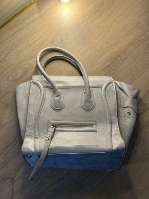 Ljusgrå handväska med dragkedja - Snygg ljusgrå handväska i konstläder med två handtag och en dragkedjeficka framtill. Väskan har en boxig form med dekorativa sömmar och metalldetaljer i guld. Perfekt för dig som gillar stilrena accessoarer med lite edge.