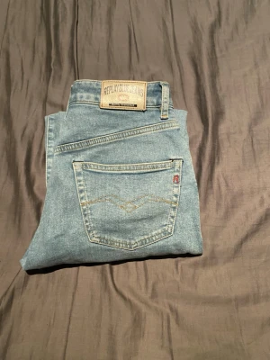 Ljusblå jeans från Replay - Säljer ett par ljusblå jeans från Replay med klassisk femficksdesign och snygga kontrastsömmar. Jeansen har en patch med Replay Blue Jeans-logga bak i midjan och är tillverkade i mjuk denim. Perfekta för en avslappnad och trendig look.