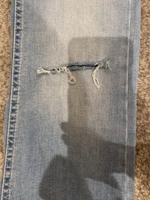 Ljusblå bootcut jeans med slitning - Säljer ett par ljusblå bootcut jeans med en snygg sliten detalj på ena benet. Jeansen har klassisk femficksdesign och är tillverkade i denim med lätt stretch. Perfekta för dig som gillar en avslappnad och trendig look.