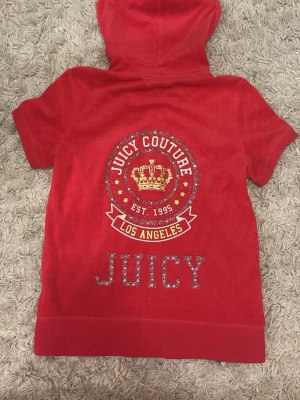 Röd Juicy Couture hoodie med dragkedja - Röd\rosa hoodie från Juicy Couture med korta ärmar, dragkedja framtill och fickor. Baksidan har ett stort tryck med krona, texten 'Juicy Couture Los Angeles' och glittriga detaljer. Tillverkad i mjuk frottéliknande material och har huva med snören. Tröjan har en liten fläck på sidan- se sista bild, går säkert bort med fläckborttagning! ♥️