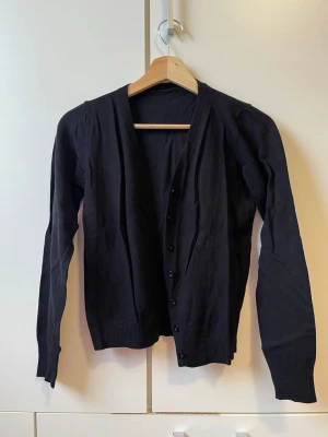 Svart basic kofta med knappar - En enkel svart kofta med långa ärmar och knäppning framtill. Koftan har ribbade muddar vid ärmslut och nederkant, vilket ger en clean och stilren look. Perfekt att slänga över en t-shirt eller linne för en avslappnad vibe. Finns inga ränder på koftan utan den är bara skrynklig på bild.