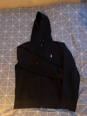 Svart hoodie från Polo Ralph Lauren - Säljer en svart hoodie från Polo Ralph Lauren med dragkedja framtill och klassisk vit logga på bröstet. Hoodien har huva med dragsko och två fickor framtill. Perfekt för dig som gillar stilrena och bekväma plagg.