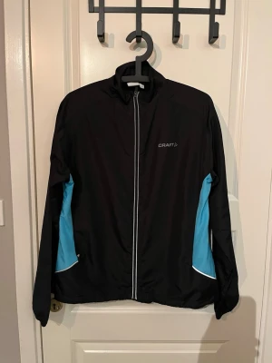 Svart träningsjacka från Craft - Sportig svart träningsjacka från Craft med ljusblå paneler på sidorna och vita detaljer. Jackan har hel dragkedja framtill, hög krage och reflekterande detaljer. Perfekt för löpning eller utomhusträning. Tillverkad i lätt och funktionellt material.