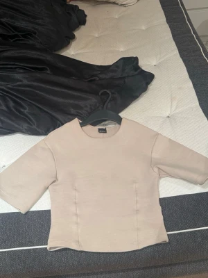 Beige topp från Gina Tricot - Säljer en stilren beige topp från Gina Tricot. Toppen har rund halsringning, raka sömmar och halvlånga ärmar. Materialet känns mjukt och stretchigt, perfekt för en clean och enkel look.