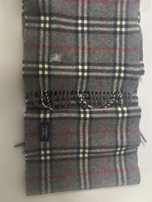 Grå rutig halsduk från Johnstons - Klassisk grå halsduk i ull från Burberry med rutigt mönster i vitt, svart och rött. Halsduken har fransar i ändarna och en mjuk, varm känsla. Perfekt accessoar för att lyfta din outfit med en tidlös look.