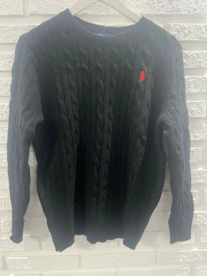 Svart kabelstickad tröja Polo Ralph Lauren - Kabelstickad svart tröja från Polo Ralph Lauren med rund halsringning och röd broderad logga på bröstet. Tröjan har långa ärmar och ribbade muddar vid ärmslut och nederkant. Perfekt för en stilren och klassisk look.