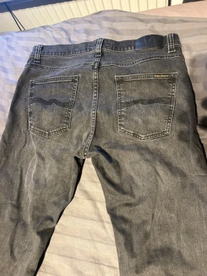 Grå/svart jeans från Nudie Jeans - Säljer ett par grå jeans från Nudie Jeans med klassisk femficksdesign och svarta detaljer på bakfickorna. Jeansen har en rak passform och är tillverkade i slitstarkt denimtyg. Perfekt för dig som gillar en avslappnad och stilren look.