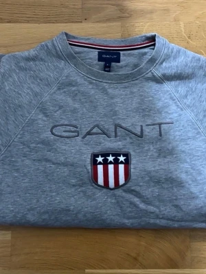 Grå sweatshirt från GANT - Snygg grå sweatshirt från GANT med broderad logga och ett emblem med stjärnor och ränder på bröstet. Tröjan har rund halsringning och långa ärmar. Tillverkad i mjuk bomull för skön känsla. Jätte bra skick. Storlek: S